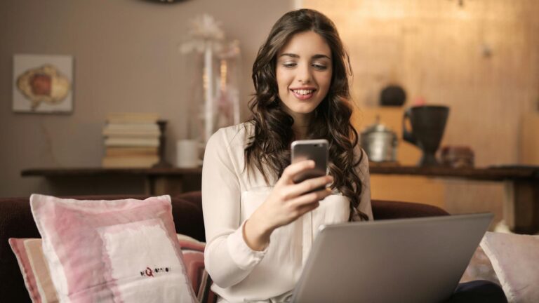 woman sitting on sofa while looking at phone with laptop on lap